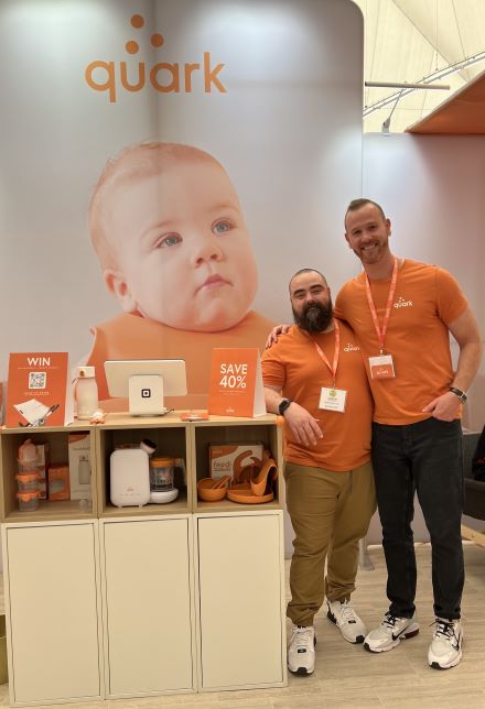 Đồng sáng lập Quark, Senez và Gurinskas đứng cạnh sản phẩm của họ tại một gian hàng triển lãm thương mại. Hình ảnh em bé ở phía sau với logo của họ.