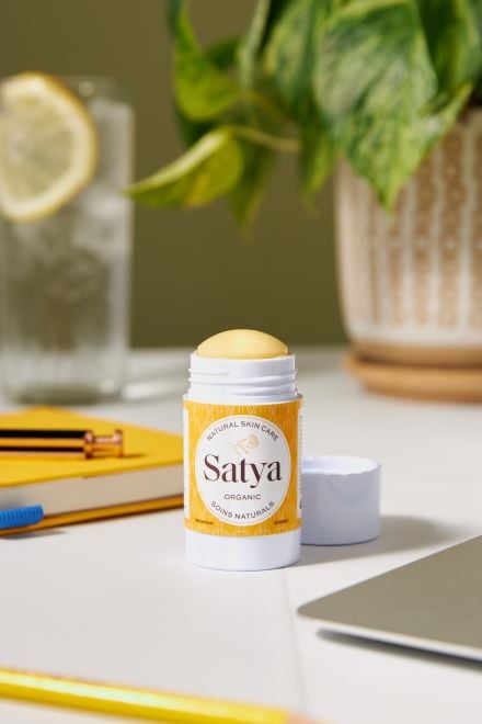 Satya product placed on a table with a glass of water and plants in the background. Product has yellow packaging and lid is open.